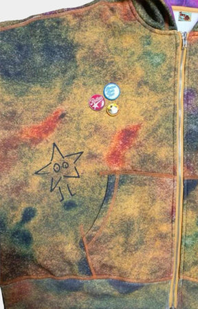 A close-up of a Dry Rot Acid Dust Zip Up Hoodie with a mottled tie-dye pattern in earthy tones of brown, yellow, and blue. The jacket features a black hand-drawn star face on the front pocket and three small colorful pins near the chest.