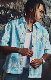 The Guapi Aqua Blue Glock 17 Leather Shirt features a striking aqua blue and white abstract pattern, a structured collar, short sleeves, distinctive toggle-style front closures, and GUAPI chest branding. Style it open over a basic tee or closed with dark denim for a bold contemporary streetwear outfit.