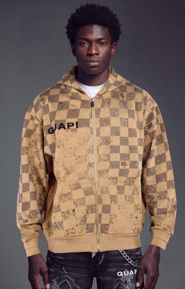 The GUAPI Desert Brown Stardust Zip-Up Hoodie features an all-over checkerboard pattern with a scattered splatter effect in a distinctive desert-brown colorway. Made from a premium cotton blend, it includes a full-zip front, adjustable hood, ribbed cuffs and hem, and GUAPI chest branding. The relaxed, modern fit makes it suitable for layering with jeans and contemporary streetwear outfits.