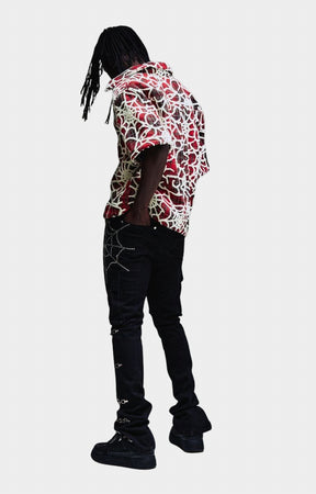 A person shown from the back wearing a short-sleeve button-up shirt with a red, white, and black abstract pattern, paired with black jeans and black shoes, against a light gray background.