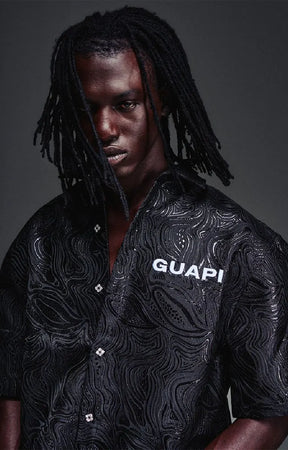 The GUAPI Liquid Black Shirt features an all-over embossed swirl pattern inspired by flowing liquid metal, subtle glossy detailing, and a tonal chest logo. Designed with a relaxed boxy fit, short sleeves, a structured collar, and custom button closures, this statement shirt brings texture and understated shine to contemporary streetwear.