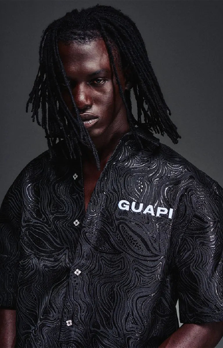 The GUAPI Liquid Black Shirt features an all-over embossed swirl pattern inspired by flowing liquid metal, subtle glossy detailing, and a tonal chest logo. Designed with a relaxed boxy fit, short sleeves, a structured collar, and custom button closures, this statement shirt brings texture and understated shine to contemporary streetwear.