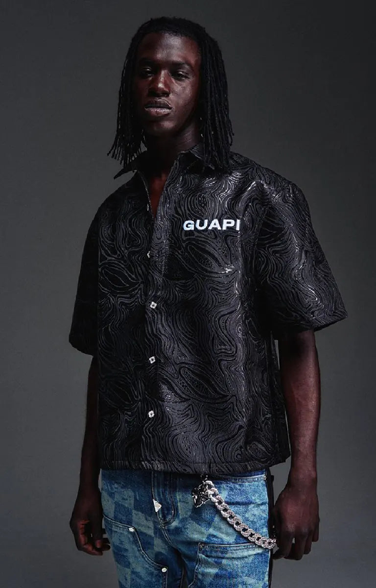 The GUAPI Liquid Black Shirt features an all-over embossed swirl pattern inspired by flowing liquid metal, subtle glossy detailing, and a tonal chest logo. Designed with a relaxed boxy fit, short sleeves, a structured collar, and custom button closures, this statement shirt brings texture and understated shine to contemporary streetwear.