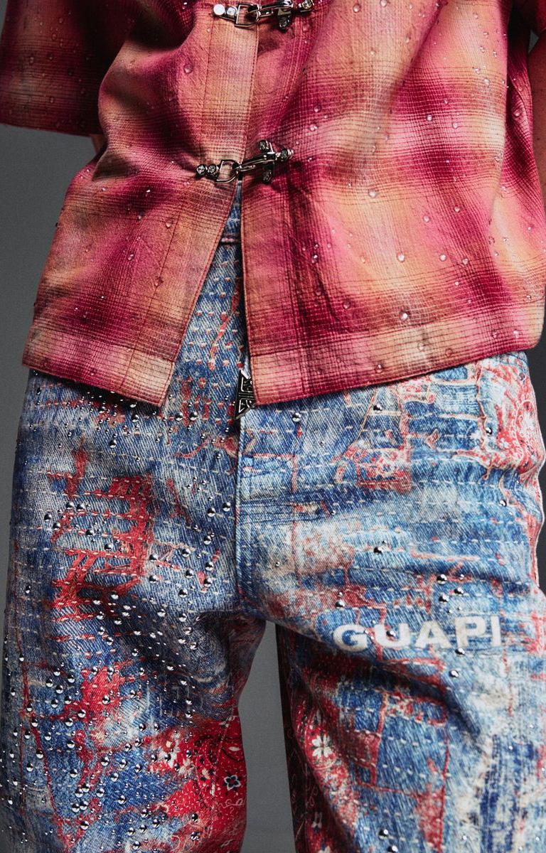 Close-up of a Guapi Red Dust Bandana Denim with an abstract multicolor print. The top is dusty rose and pink with dark metal toggle closures, while the pants are blue with red splatter-like patterns and silver rhinestone embellishments.