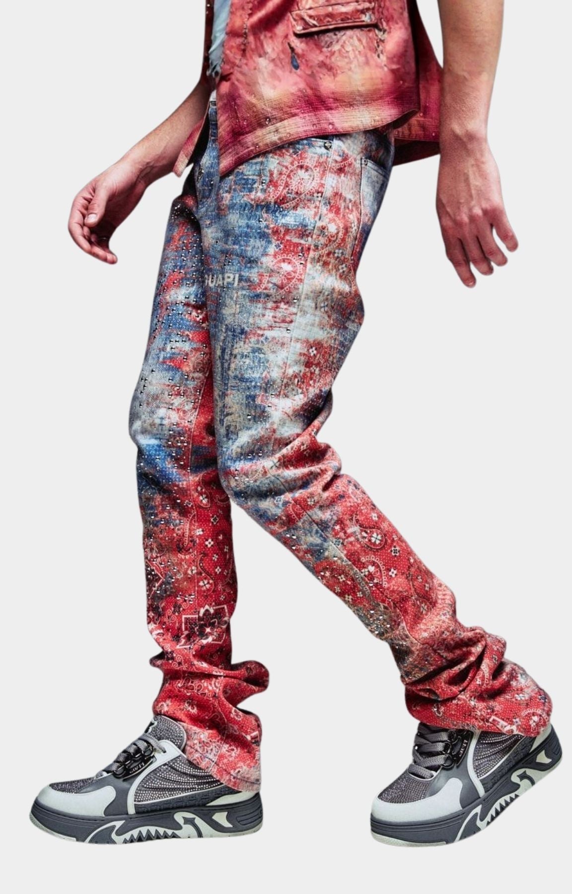 Guapi Red Dust Bandana Denim distressed, faded red and blue jeans with a tie-dye pattern, paired with gray and white sneakers.