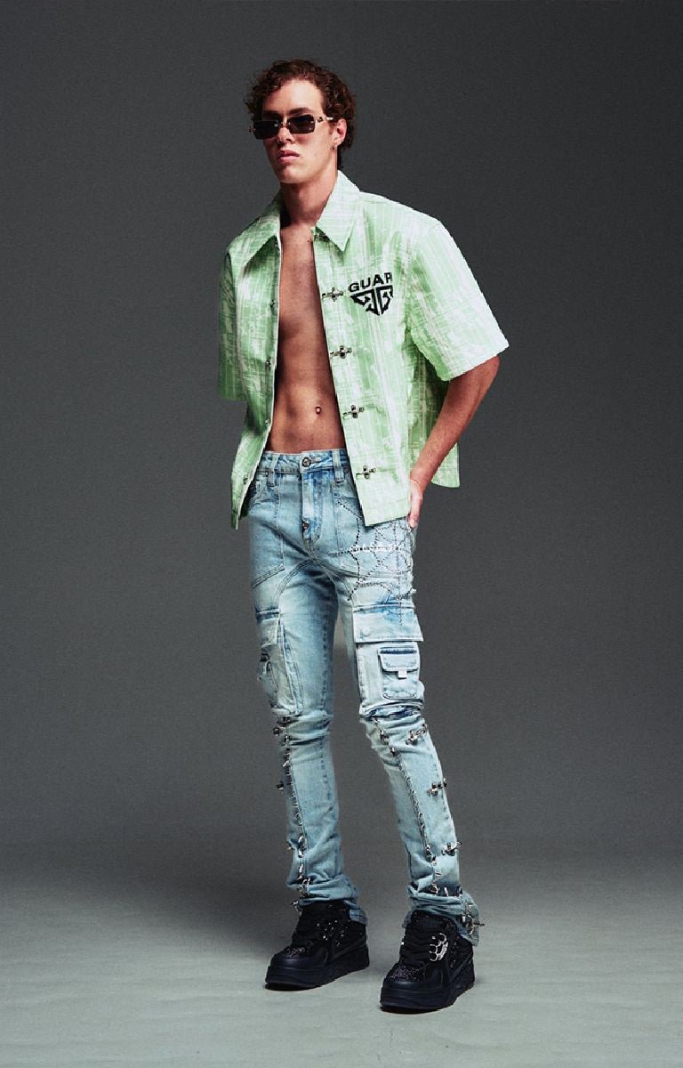 A male model wearing Guapi light green short-sleeve button-up shirt with a black Gucci logo on the chest, aqua blue light-wash distressed jeans, and black sneakers.