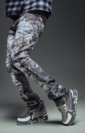 Guapi Hakai Spider stacked denim jeans with slim streetwear fit, distressed, graffiti-style black and white patterned jogger pants with gray and white high-top sneakers.