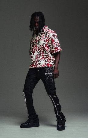 A model wearing a guapi blood red short-sleeve button-up shirt with a red, white, and black abstract pattern, paired with guapi obsidian black cargo pants featuring spiderweb graphics and black platform sneakers.