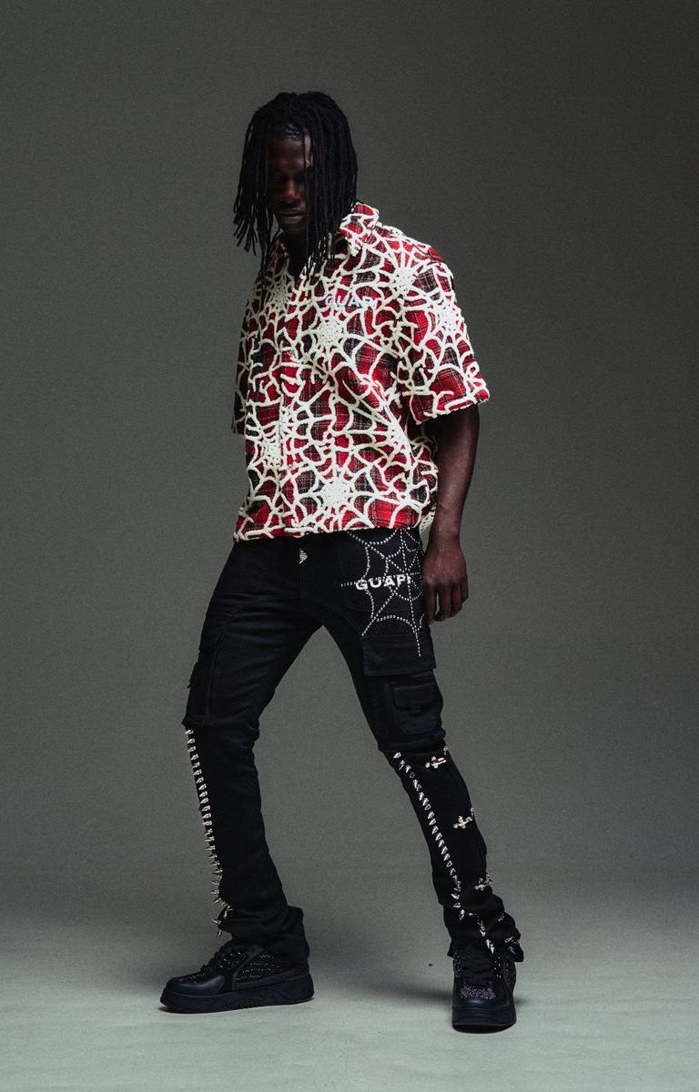 A model wearing a guapi blood red short-sleeve button-up shirt with a red, white, and black abstract pattern, paired with guapi obsidian black cargo pants featuring spiderweb graphics and black platform sneakers.