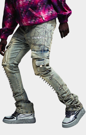 Guapi Vintage Spikes Cargo Denim in faded grey wash heavily distressed and embellished gray cargo-style jeans with metal studs, chain details, and ripped sections, paired with a purple patterned top and silver sneakers. 