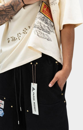 A close-up of a person wearing an oversized cream paper money graphic T-shirt and paper money black shorts with gold-tone hardware and a visible Paper Money hangtag.