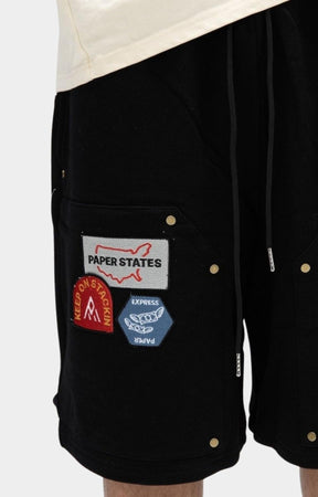 Close-up of black shorts with a front pocket decorated with multiple colorful patches, including a white 'PAPER STATES' patch and a red circular patch, shown on a model.