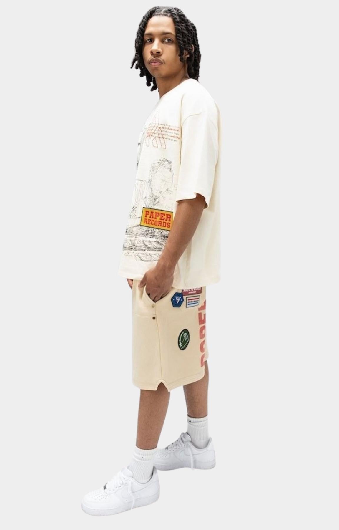 Angled view of Paper Money Paper States oversized graphic cream-colored graphic t-shirt and matching graphic shorts, paired with white socks and white sneakers.