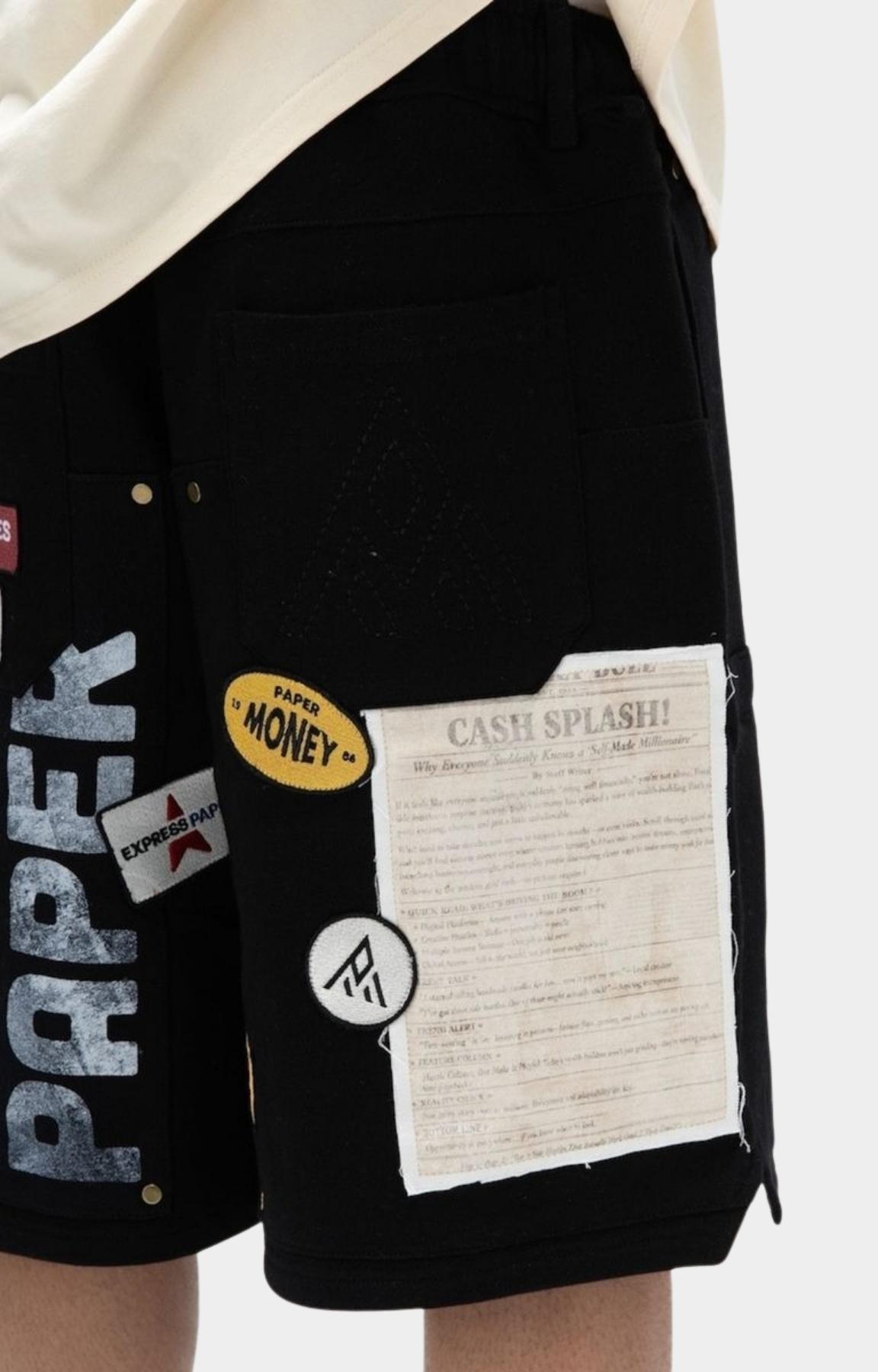 Close-up of paper money black shorts with multiple graphic patches and printed details, worn with a cream shirt. Visible patches include a yellow 'MONEY' sticker, a white 'CASE SPLASH!' note patch, and a circular logo patch.