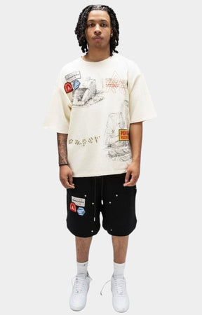 A model wearing Paper Money Paper States cream graphic tee for premium streetwear styling with black shorts. The t-shirt features a detailed astronaut illustration and graphic patches, while the shorts also have small graphic details.