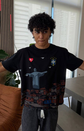 Model wearing  Red Letters Favelas black graphic t-shirt featuring a stylized depiction of Christ the Redeemer and Rio de Janeiro rooftops, with a red heart logo and white birds on the chest, paired with gray jean. 