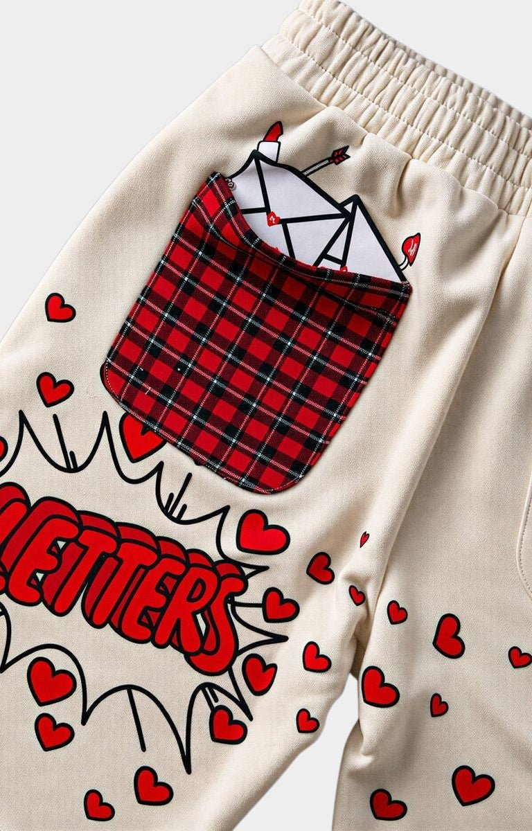 Close-up of cream-colored red letters shorts featuring a red and black plaid pocket graphic with a white envelope, red comic-style hearts, and the word 'LETTERS' in red comic lettering.