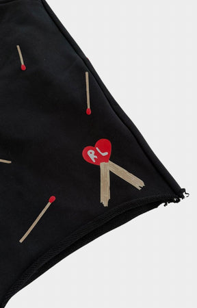 Close-up of red letters black shorts, featuring a pattern of wooden matchsticks with red tips and a central red heart patch with the letters 'RL' in white.
