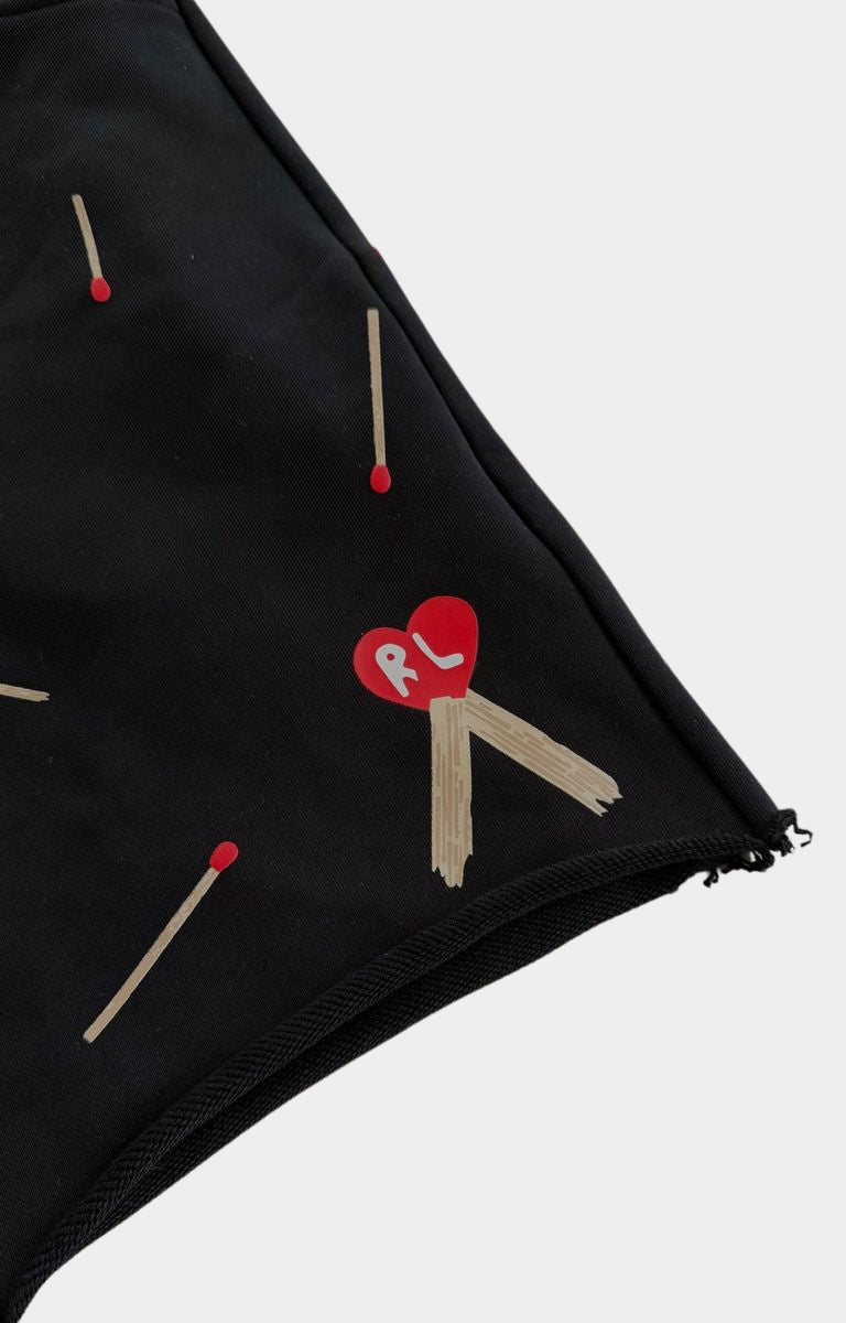 Close-up of red letters black shorts, featuring a pattern of wooden matchsticks with red tips and a central red heart patch with the letters 'RL' in white.