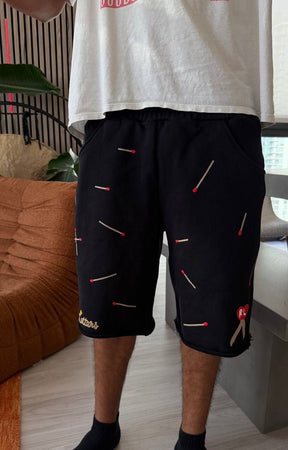 Model wearing Red Letters black shorts with embroidered matchstick patterns in red, white, and black at front. 