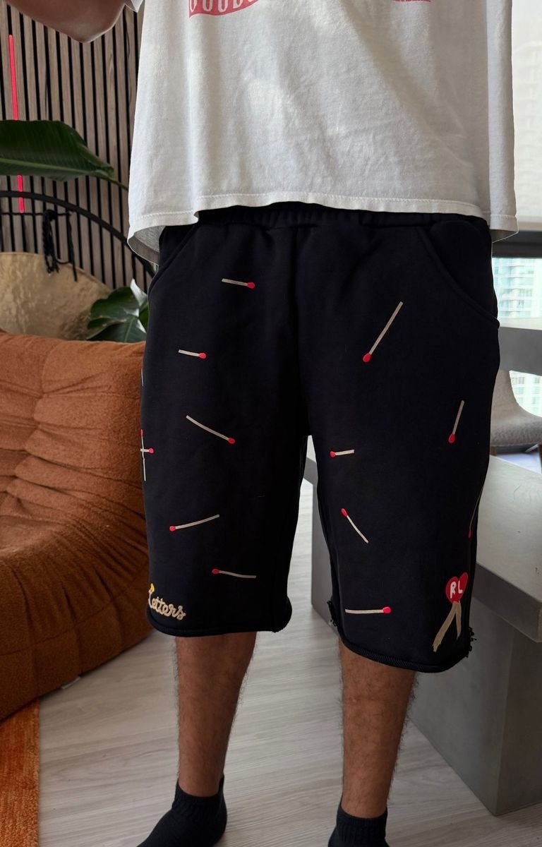 Model wearing Red Letters black shorts with embroidered matchstick patterns in red, white, and black at front. 