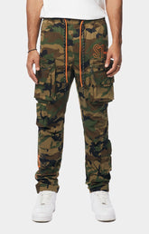 Smoke Rise camouflage cargo jogger pants with multiple flap pockets, an orange drawstring, and an orange embroidered logo on the thigh, paired with white sneakers.