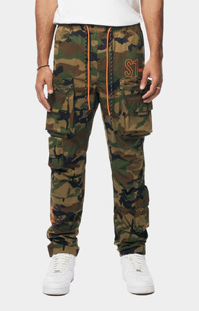 Smoke Rise camouflage cargo jogger pants with multiple flap pockets, an orange drawstring, and an orange embroidered logo on the thigh, paired with white sneakers.