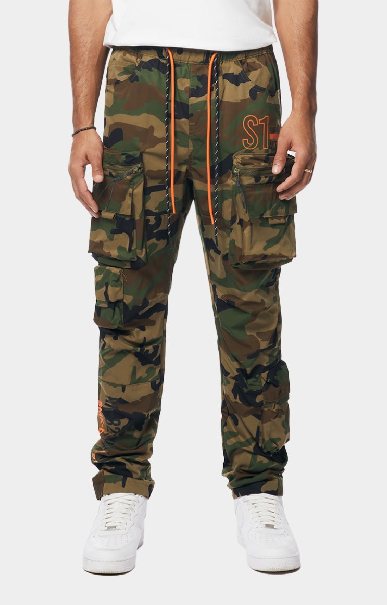 Smoke Rise camouflage cargo jogger pants with multiple flap pockets, an orange drawstring, and an orange embroidered logo on the thigh, paired with white sneakers.